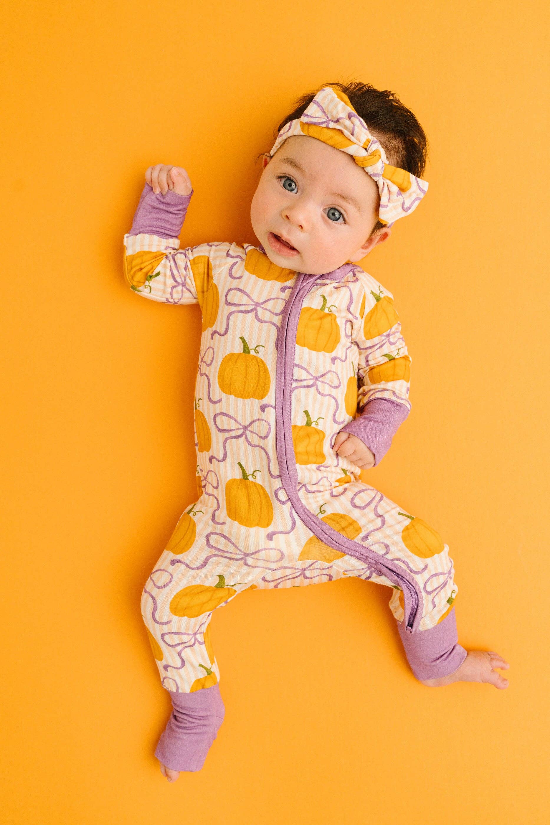 A baby lying on their back against a solid orange background, wearing a cream-colored footed sleeper with a yellow pumpkin and purple bow pattern, matching purple cuffs, and a matching purple and yellow headband.