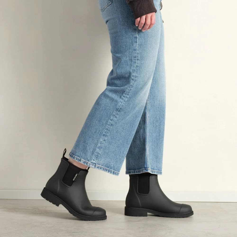 Person wearing black rubber rain boots and blue jeans on a neutral background
