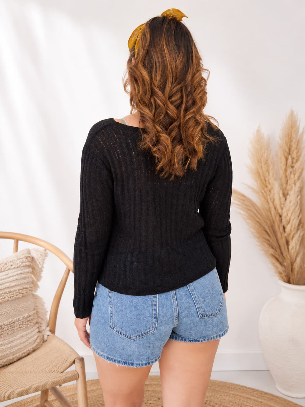Woman wearing a black sweater and denim shorts with a yellow headband, standing in a room with a chair and decorative elements.