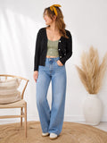 Woman wearing a black cardigan, green top, and blue jeans in a room with a chair and decorative elements.