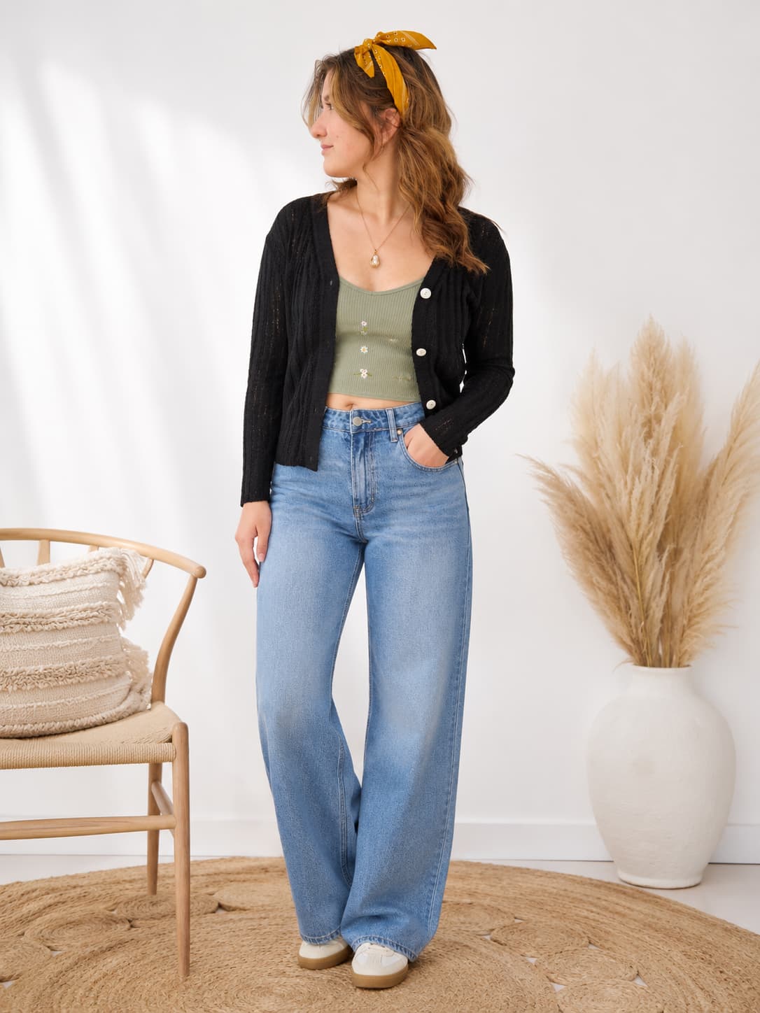 Woman wearing a black cardigan, green top, and blue jeans in a room with a chair and decorative elements.