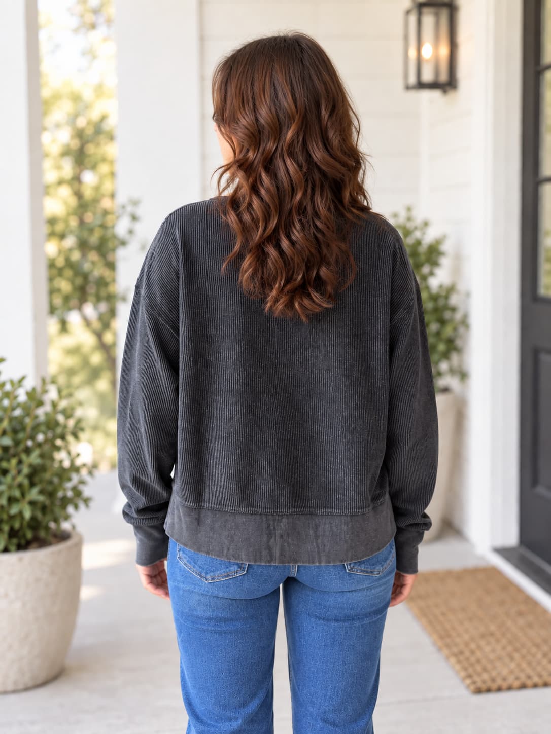 A woman viewed from behind wearing a dark charcoal gray washed sweatshirt and blue jeans, standing on a front porch with white columns and plants.