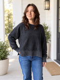 A woman wearing a dark charcoal gray washed crewneck sweatshirt and blue jeans, standing on a white porch with potted plants.