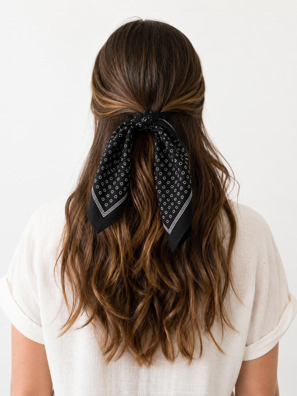 A woman viewed from behind wearing a black square scarf with a white daisy pattern tied in a loose bow at the base of her hair, which is styled with light brown highlights.