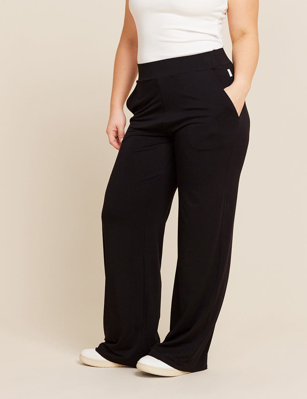 A model wearing black wide-leg trousers with a white sleeveless top and white sneakers, standing against a solid beige studio background.