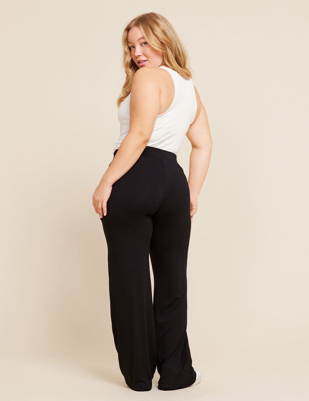 A woman modeling a pair of black wide-leg pants with a white tank top, shown from a three-quarters back view against a solid beige studio background.