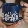 A close-up of a person holding a glossy cobalt blue ceramic mug with a white floral pattern and a beige base, showing their hands with manicured nails.
