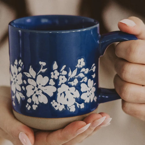 A close-up of a person holding a glossy cobalt blue ceramic mug with a white floral pattern and a beige base, showing their hands with manicured nails.