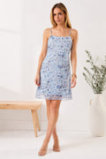 Woman wearing a light blue floral dress standing in a minimalistic room with a plant and wooden bench.