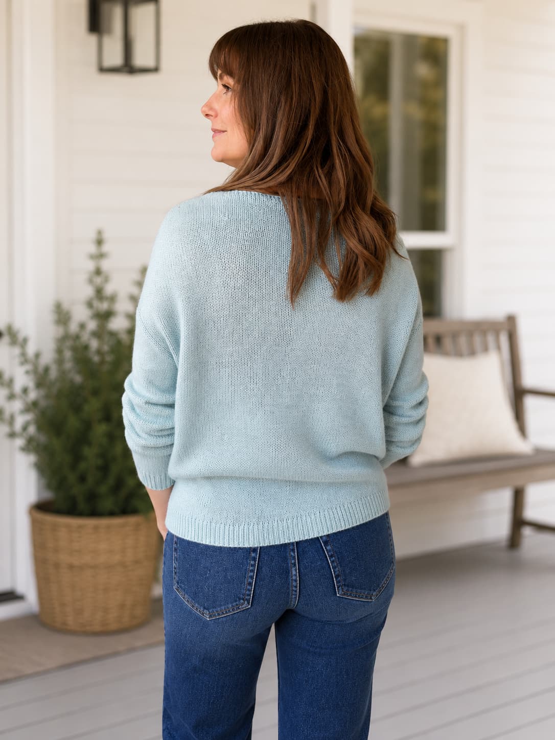 Back view of a  woman wearing a light blue knit sweater and blue jeans, photographed from the back on a porch with a bench and potted plant in the background.