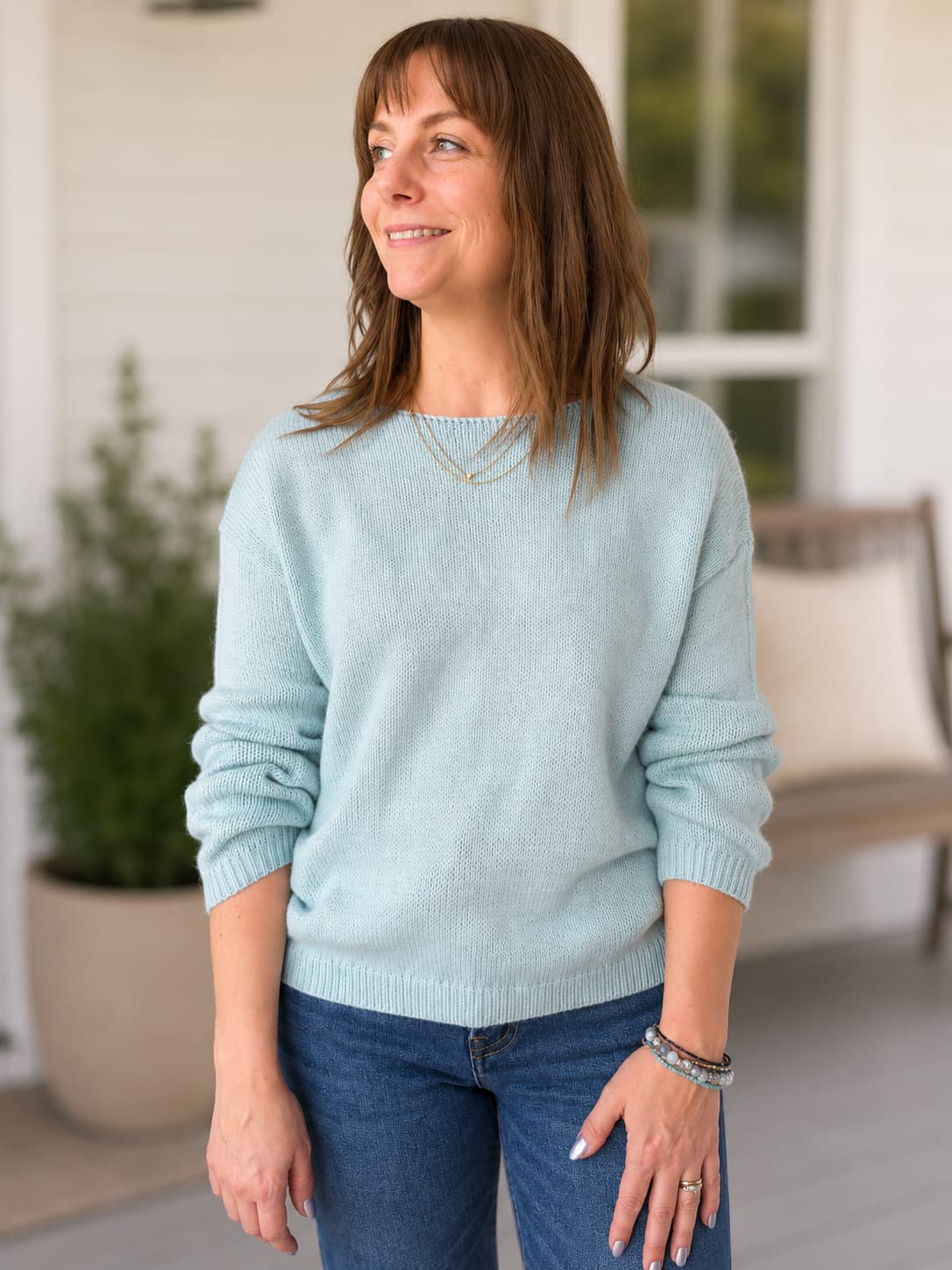 A woman wearing a super soft light weight blue knit crewneck sweater and blue jeans, standing on a porch with a potted plant and a bench in the background.