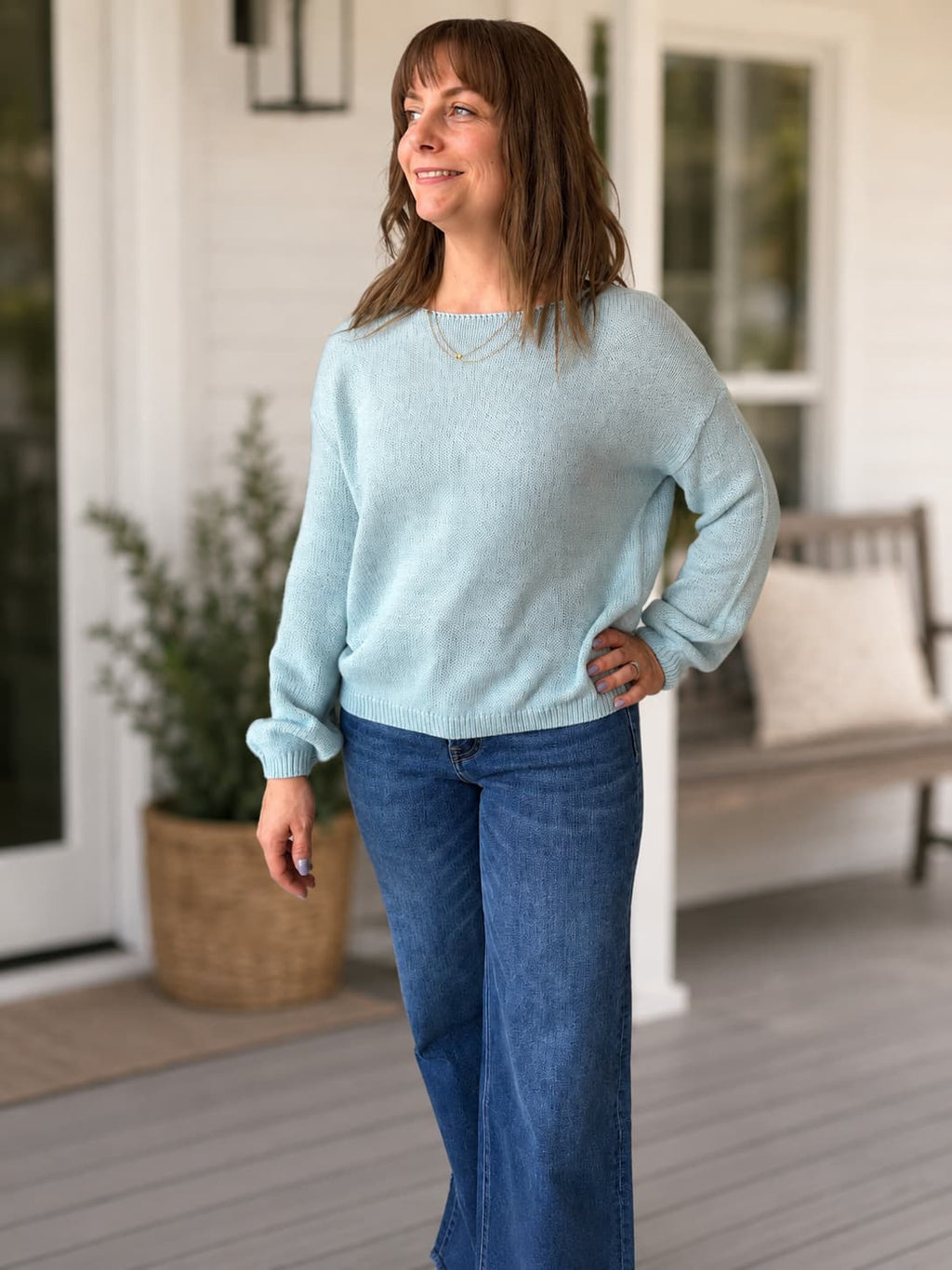 A woman wearing a soft light weight, light blue knit sweater and blue jeans stands on a porch, looking to the side. 
