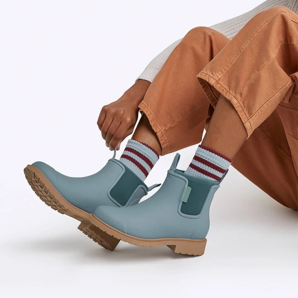 Light blue rain boots with brown soles worn by a person in tan pants and striped socks.