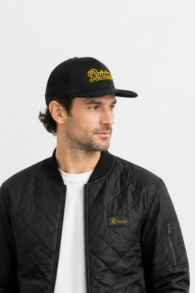 A man wearing a black quilted bomber jacket, khaki pants, a white t-shirt, black and white sneakers, and a black baseball cap with yellow embroidery, standing in a bright room with a plant and wooden bench.