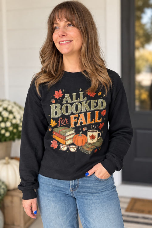 A woman wearing a black long-sleeve crewneck sweatshirt with a colorful autumn-themed graphic that reads 'ALL BOOKED FOR FALL' with books, a pumpkin, and a mug. She is wearing blue jeans and standing in front of a white house door.