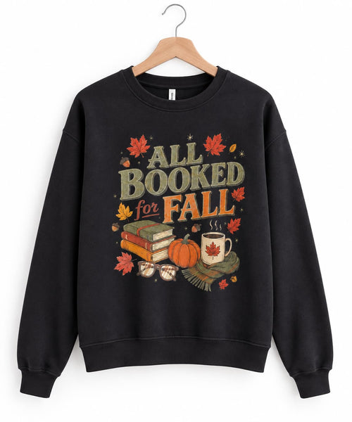 A black crewneck sweatshirt hanging on a wooden hanger against a white background, featuring a colorful graphic print that reads 'ALL BOOKED for FALL' with autumn-themed illustrations including leaves, a pumpkin, books, and a mug.