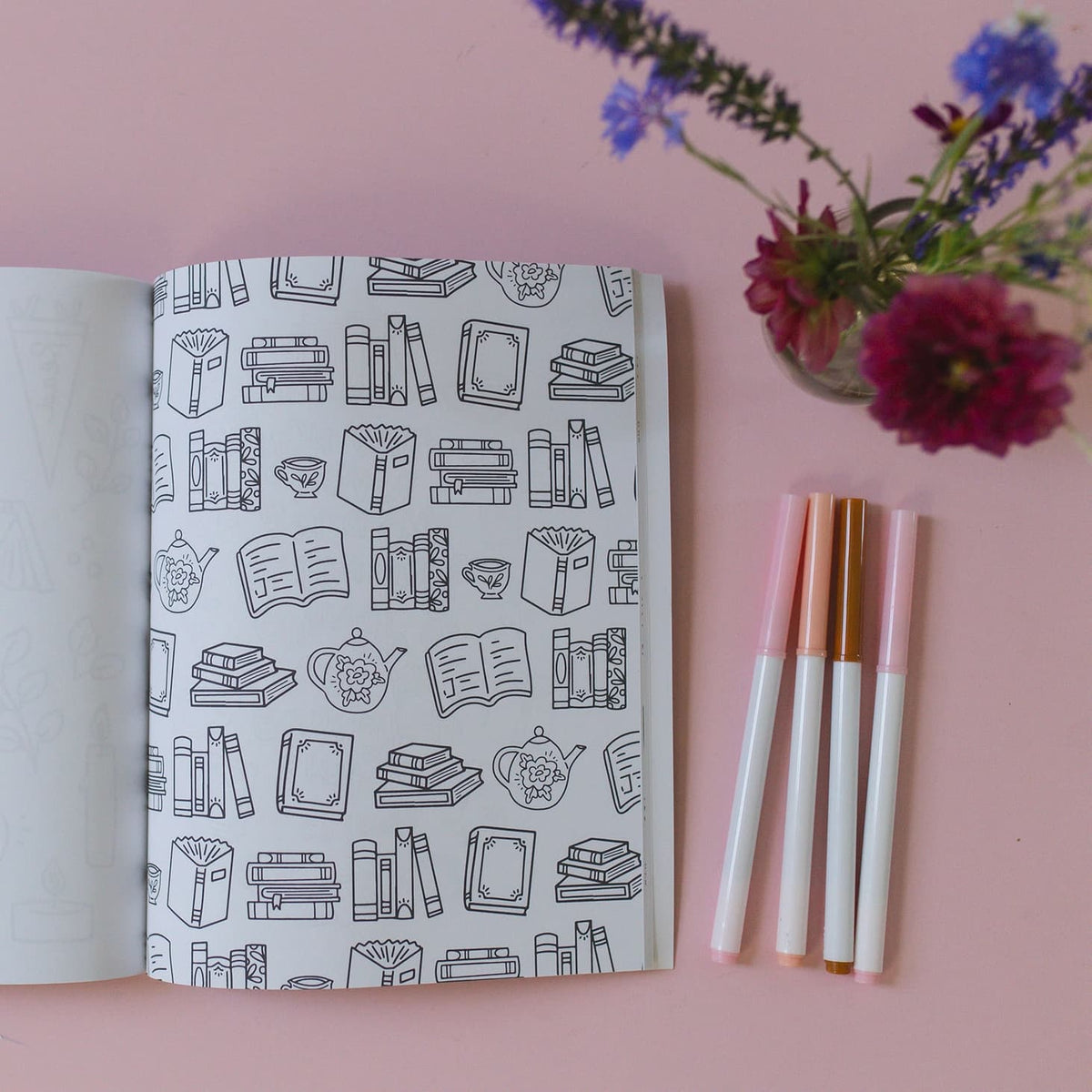 Notebook with book-themed cover, markers, and flowers on a pink background