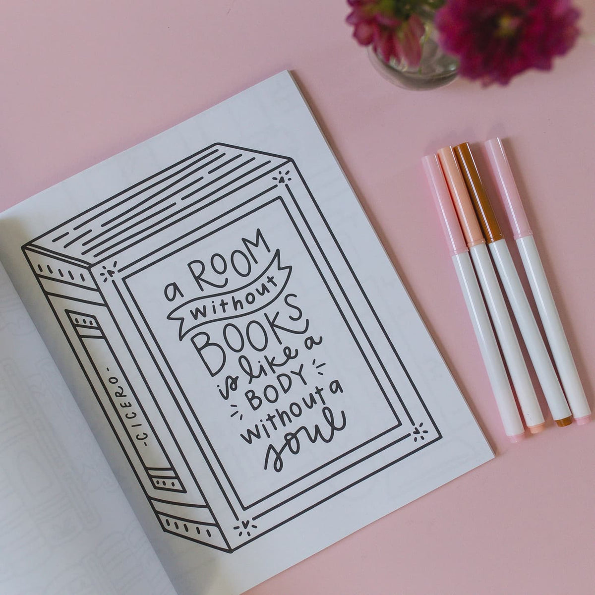 Open book with a quote about books on a pink background with markers and flowers.