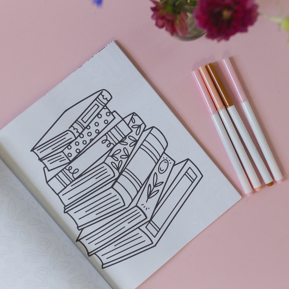 A black line art illustration of a stack of books on a white notebook page, styled on a pink background with four pastel pink and rose gold pens and flowers.