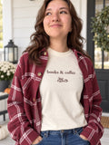 A woman wearing a cream crewneck t-shirt with 'books & coffee' and a small coffee cup graphic printed on the chest, layered under a maroon and white plaid button-down flannel shirt, paired with blue jeans.