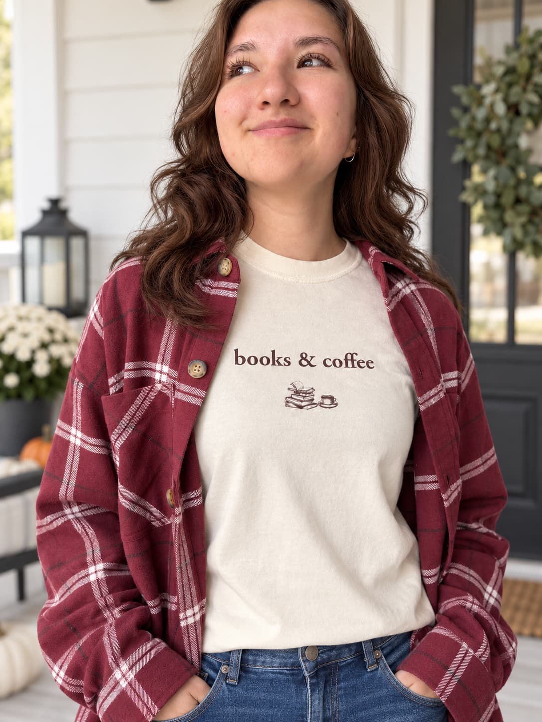 A woman wearing a cream crewneck t-shirt with 'books & coffee' and a small coffee cup graphic printed on the chest, layered under a maroon and white plaid button-down flannel shirt, paired with blue jeans.