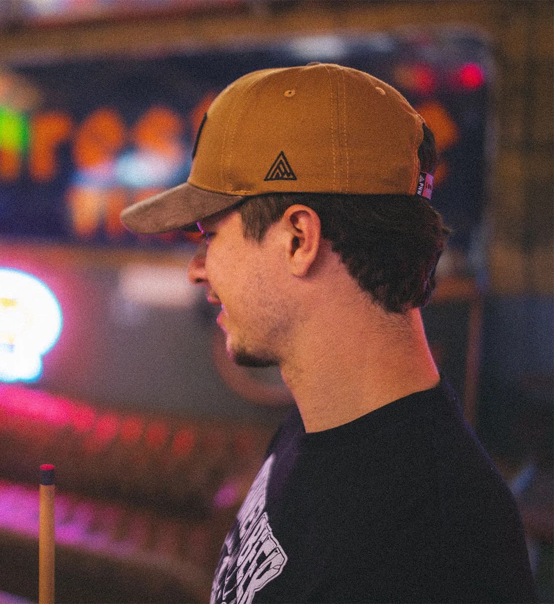 A man in profile wearing a tan baseball cap with a black triangular logo and a black t-shirt, photographed in a dimly lit, colorful indoor environment.