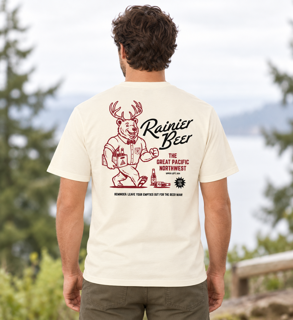 A man viewed from behind wearing a cream-colored t-shirt with a large red graphic on the back. The graphic features a deer mascot holding beer glasses and the text 'Rainier Beer'.