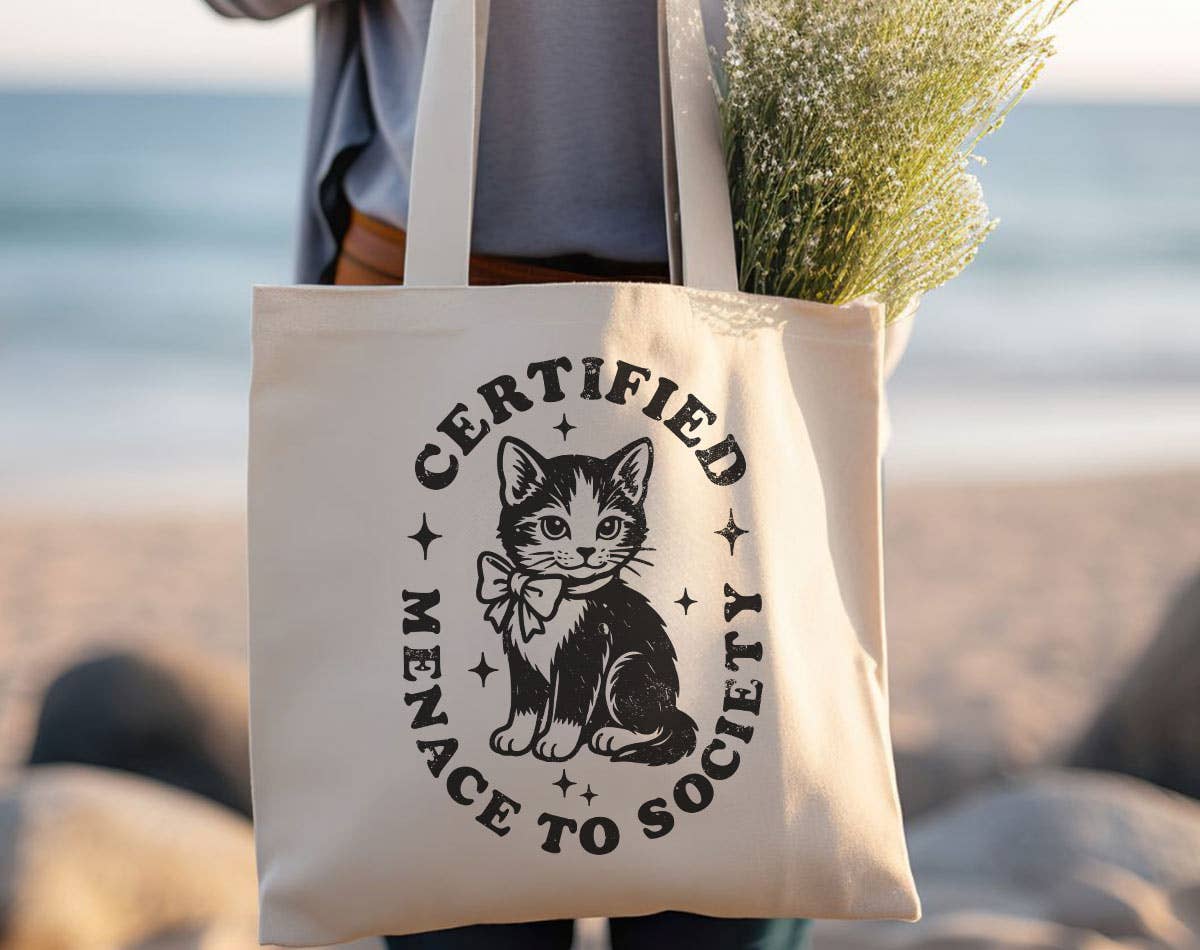 Tote bag with a cat design and text held by a person on a beach.
