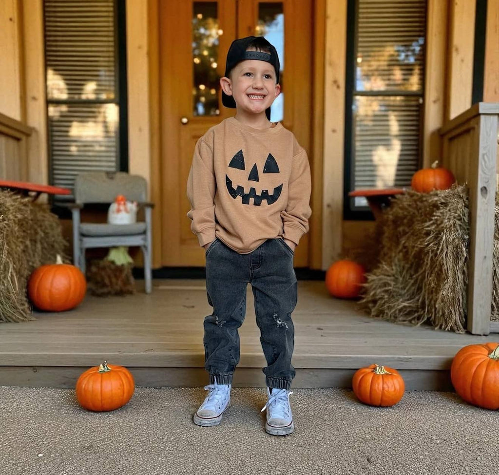 A young boy stands on a wooden porch decorated with pumpkins and hay bales, wearing a tan crewneck sweatshirt with a black jack-o'-lantern face graphic, dark gray pants, and a black baseball cap.