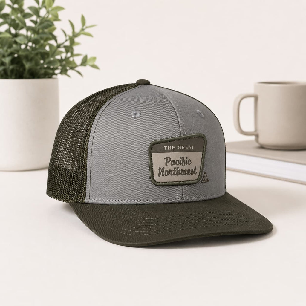 A gray and dark olive green trucker cap with a mesh back, featuring a rectangular patch on the front that reads 'THE GREAT Pacific Northwest'. The cap is styled on a white surface with a potted plant and a mug in the background.