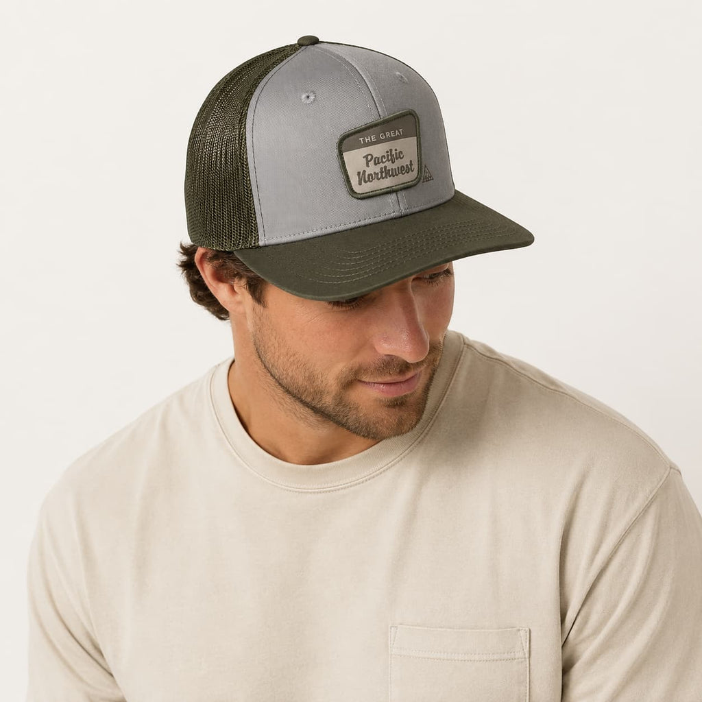 A man wearing a gray and olive green Pacific Handcrafted trucker hat with a leather patch, paired with a beige crewneck shirt against a light off-white background.