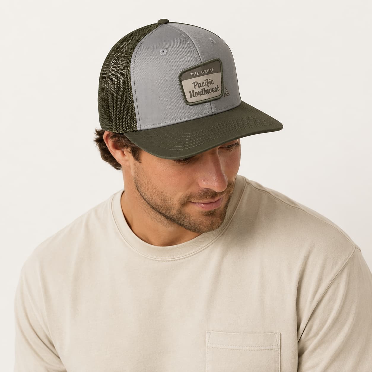 A man wearing a gray and olive green Pacific Handcrafted trucker hat with a leather patch, paired with a beige crewneck shirt against a light off-white background.
