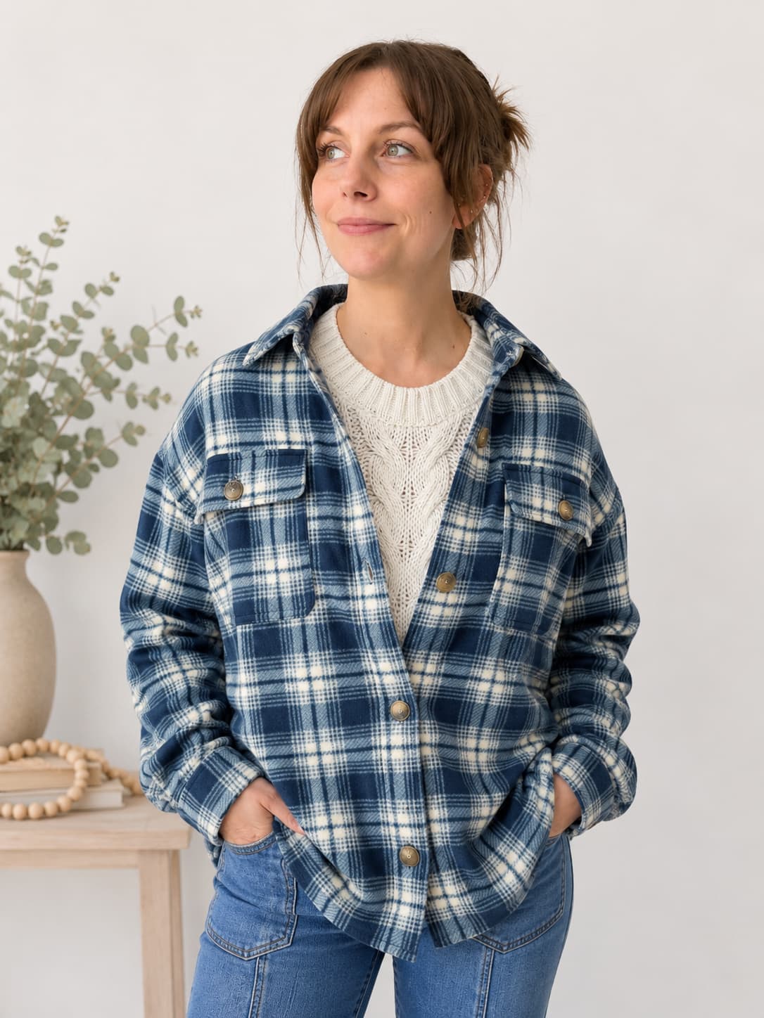 A woman wearing a blue and white plaid flannel button-up shirt jacket over a cream knit sweater and blue jeans, standing in a brightly lit indoor setting.