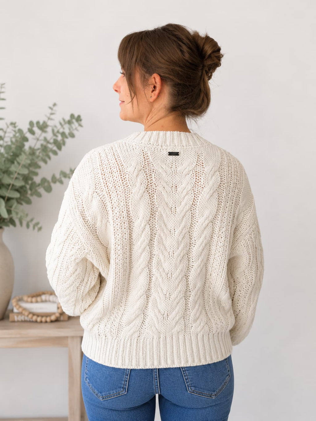 A woman viewed from behind wearing a cream cable-knit sweater and blue jeans, standing in a bright indoor setting with a light wall and a small table with greenery on the left.