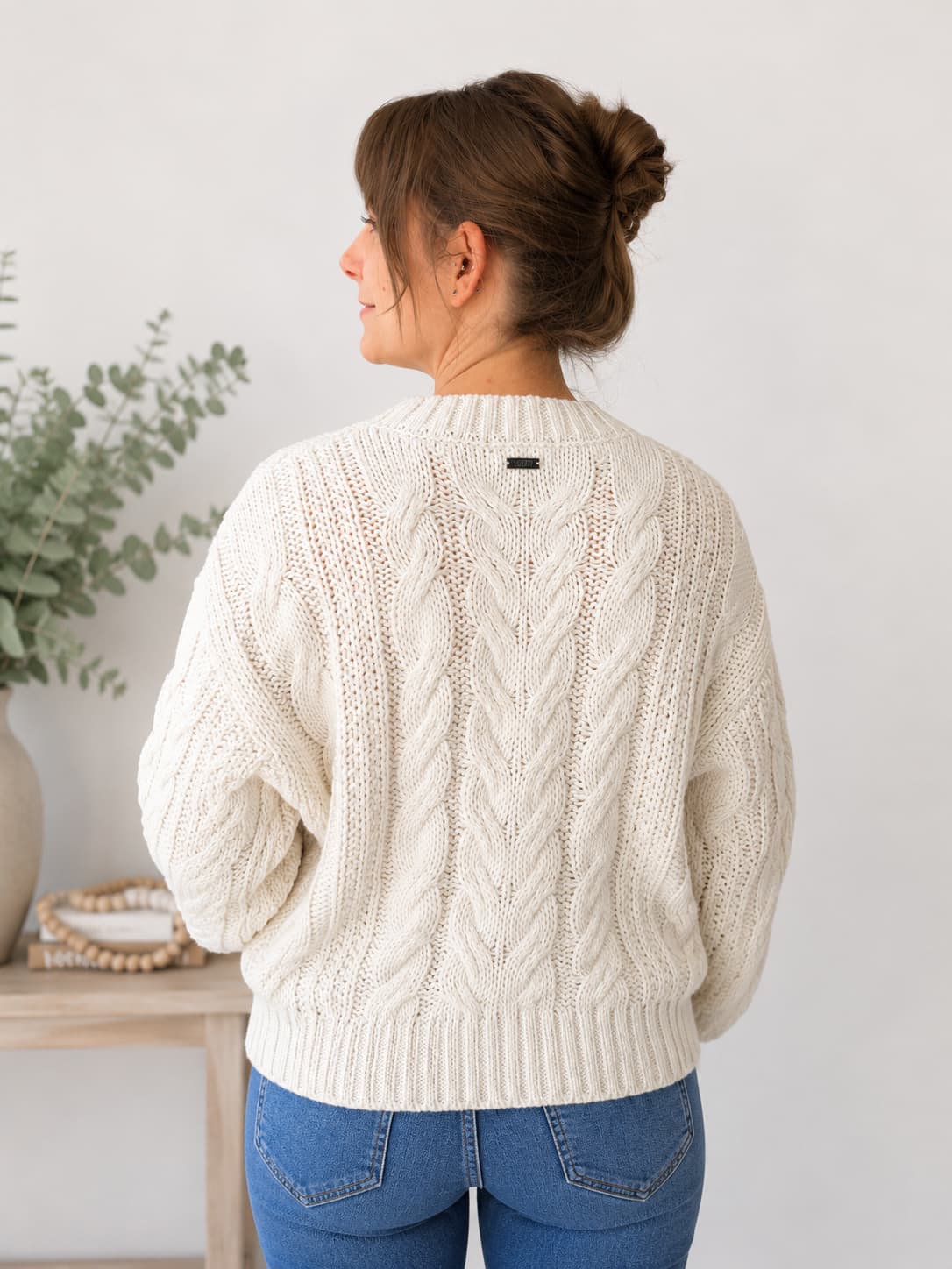 A woman viewed from behind wearing a cream cable-knit sweater and blue jeans, standing in a bright indoor setting with a light wall and a small table with greenery on the left.