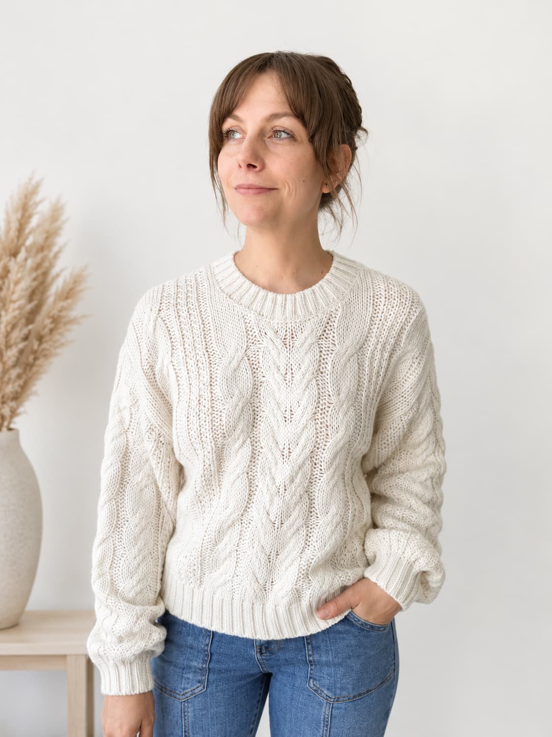 A woman wearing a cream cable-knit crewneck sweater and blue jeans, standing against a light gray wall with a vase of dried grass on a side table.