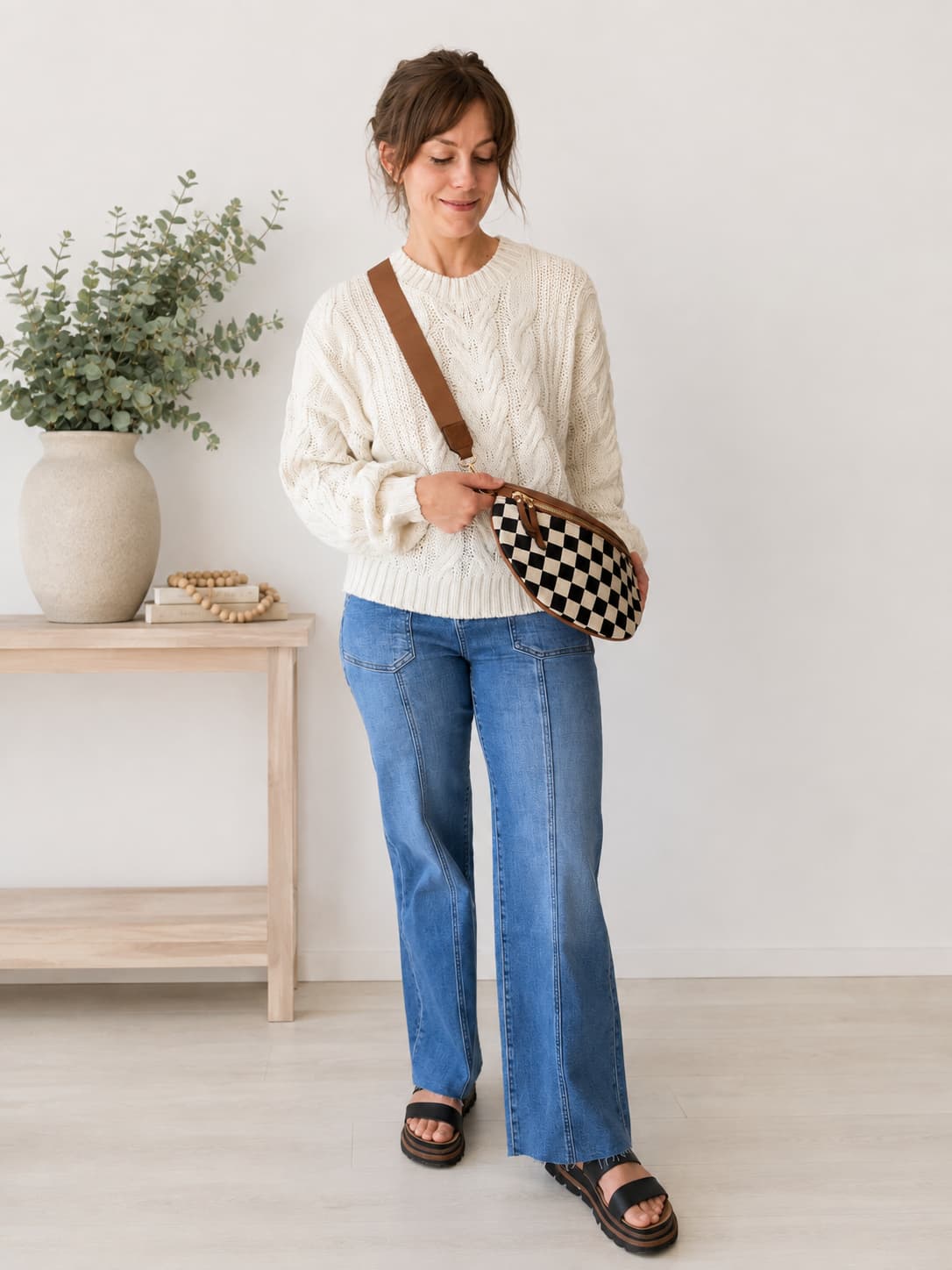 A woman stands in a bright room wearing a cream cable-knit sweater, blue jeans, and black sandals, while carrying a brown leather crossbody bag with a black and white checkered pattern.