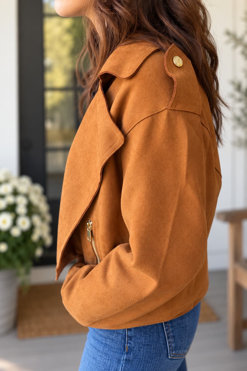 A woman wearing a camel brown suede moto jacket with gold-tone hardware, paired with blue jeans, shown from a side profile in a bright, modern indoor setting.