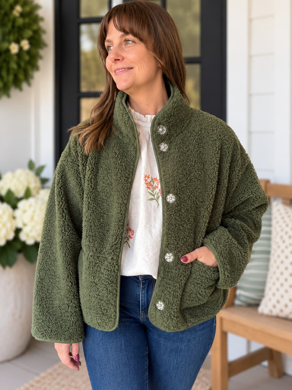 A woman wearing an olive green sherpa fleece button-front jacket with silver buttons, layered over a white floral top and blue jeans, standing on a porch.