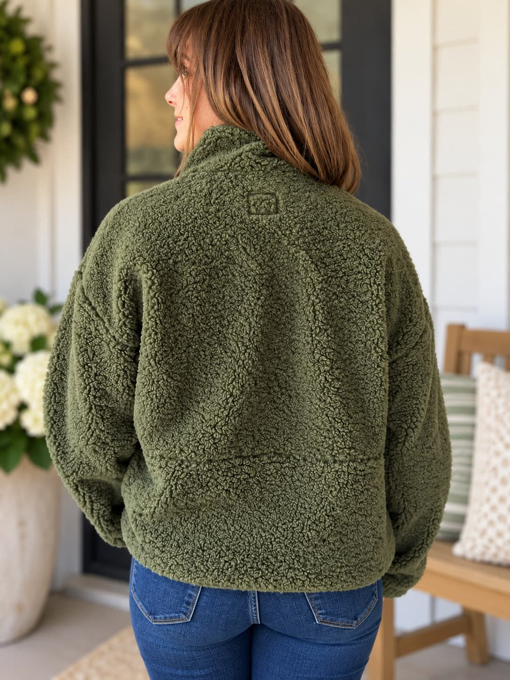 A woman viewed from the back wearing an olive green sherpa fleece jacket and blue jeans, standing on a porch.