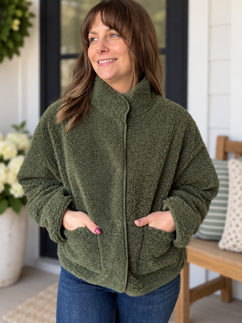 A woman wearing a cozy, textured olive green fleece jacket with a stand-up collar and front pockets, paired with blue jeans, standing on a porch.