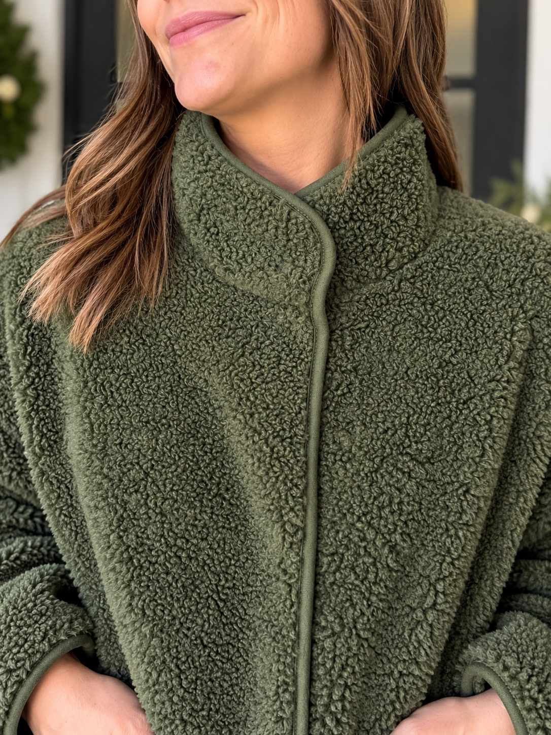 A close-up of a woman wearing an olive green sherpa fleece jacket with a high collar and front zipper.