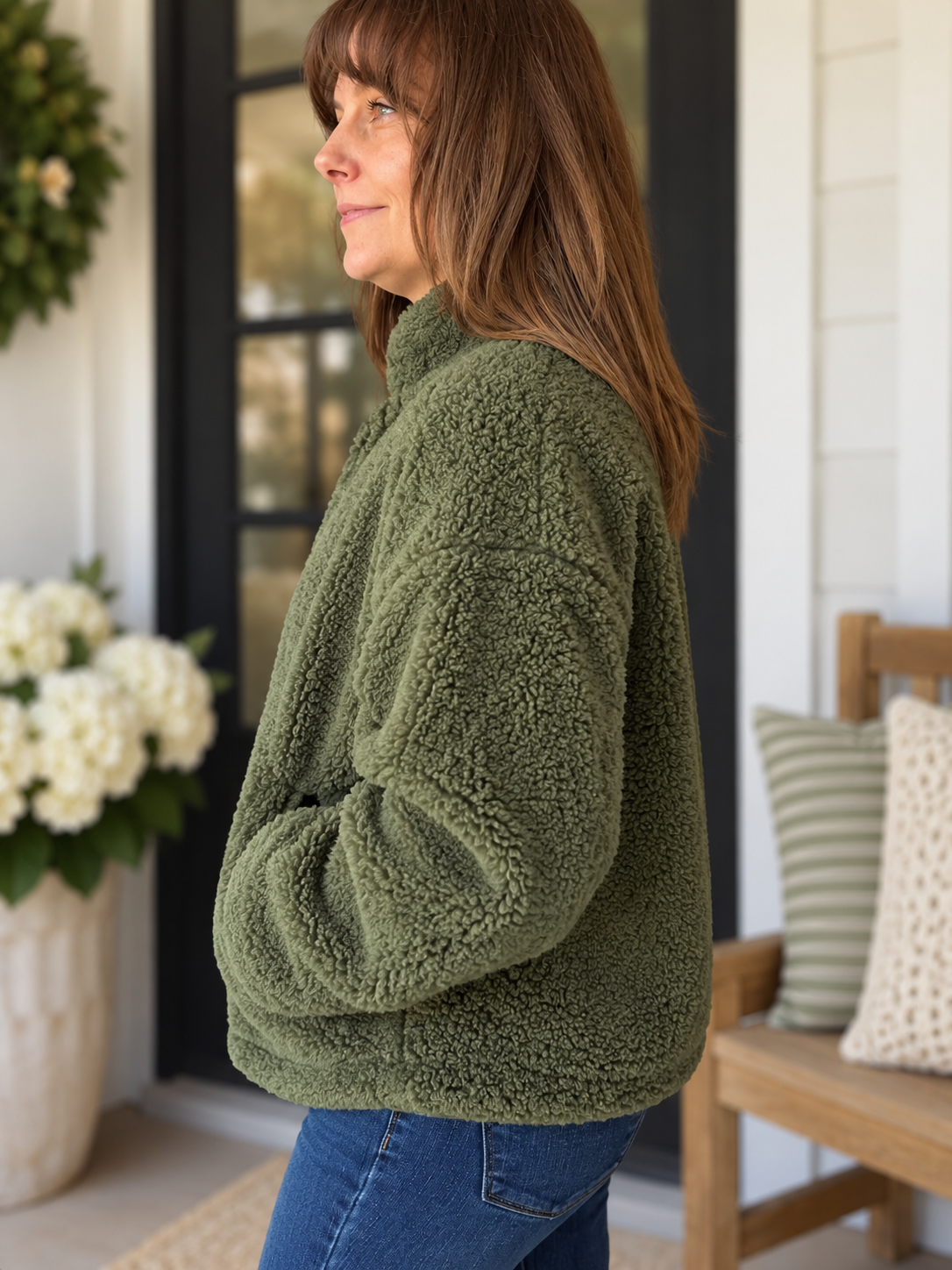 A woman wearing an olive green fleece pullover sweater and blue jeans, standing on a porch next to a wooden bench with pillows.