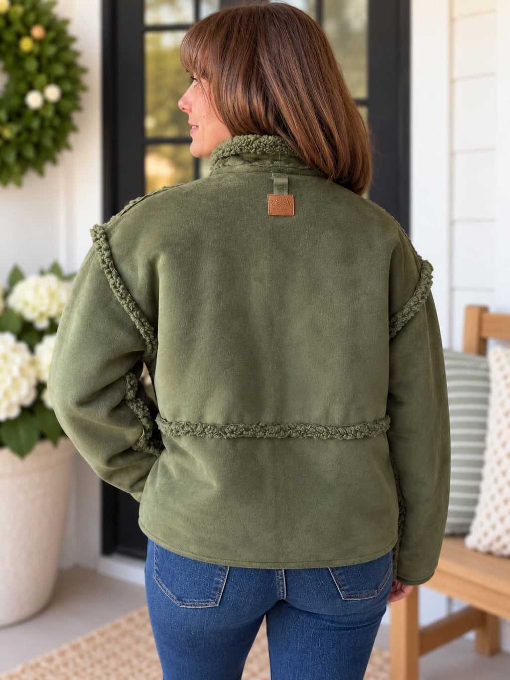 A woman viewed from the back wearing an olive green fleece jacket with textured trim details, paired with blue jeans, standing on a porch.