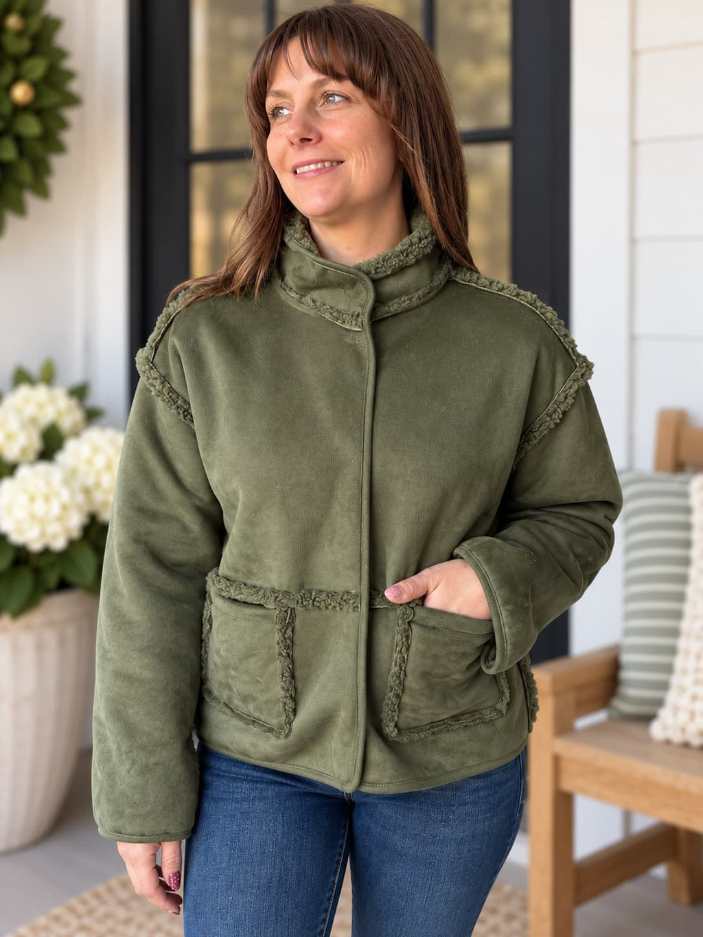 A woman wearing an olive green fleece jacket with fringed trim details, paired with blue jeans, standing on a porch.