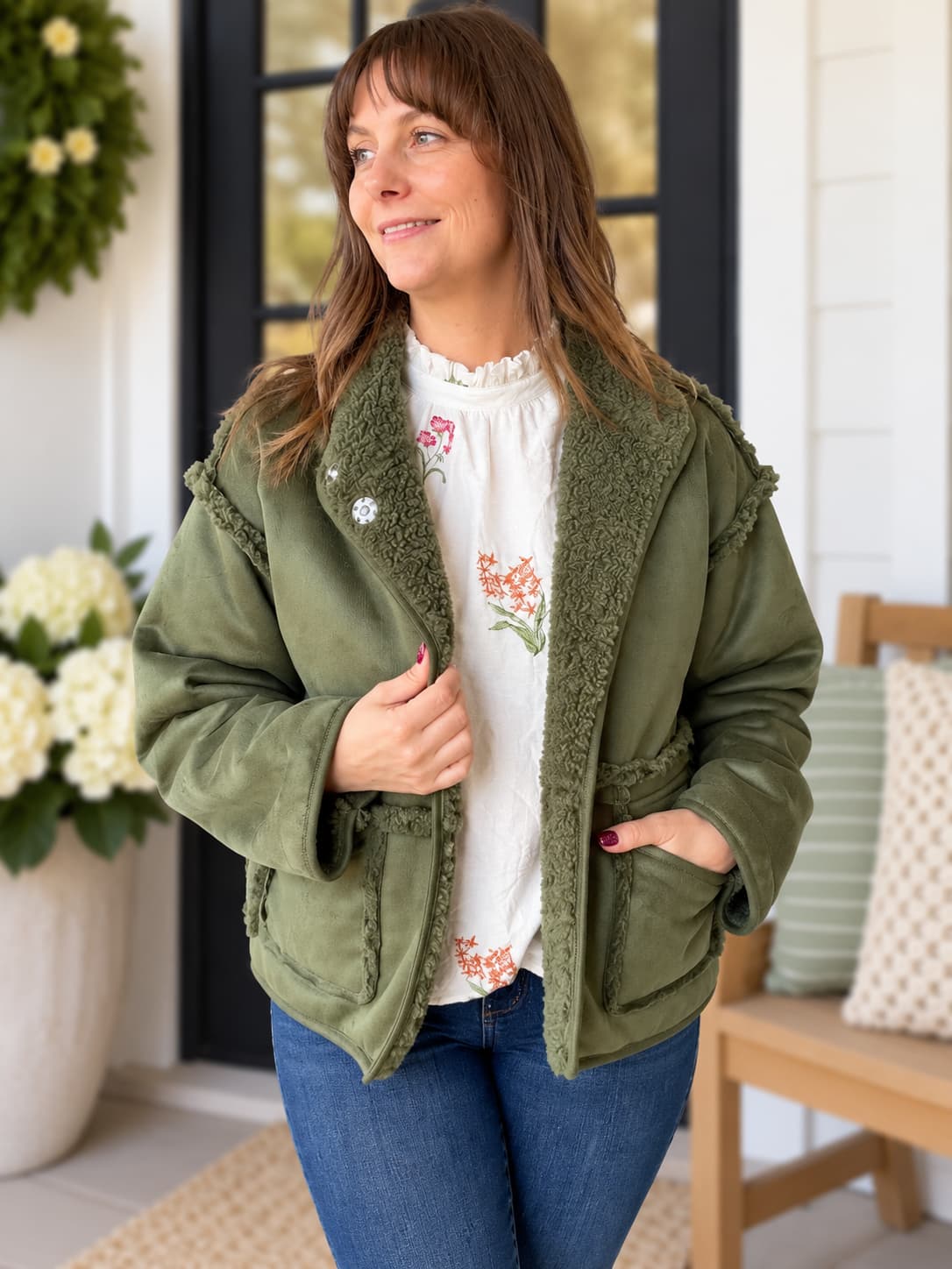 A woman wearing an olive green fleece-lined jacket over a white floral top and blue jeans, standing on a porch with a wooden bench and potted flowers in the background.