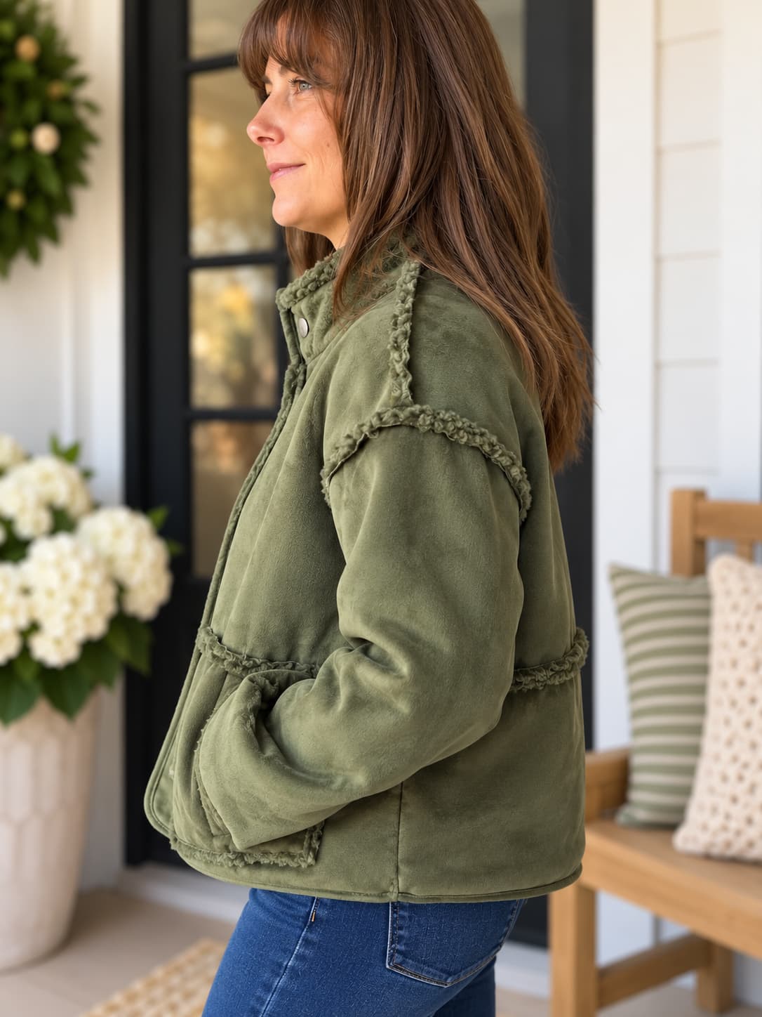 A woman wearing an olive green fleece jacket with decorative braided trim, standing on a porch next to a wooden bench with pillows.