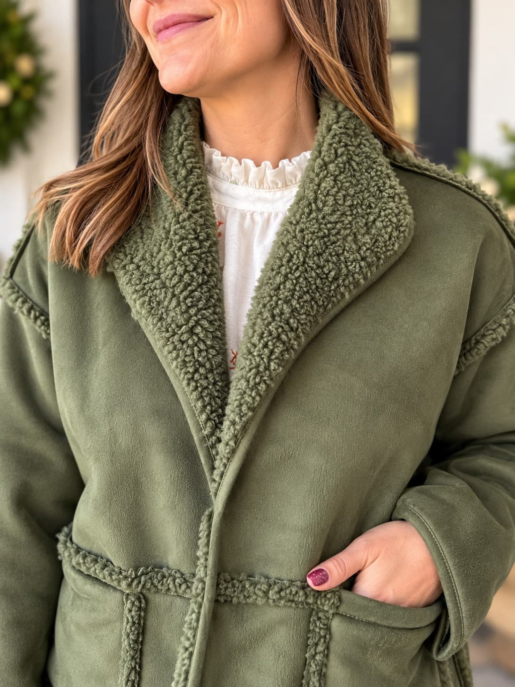 A close-up of a woman wearing an olive green suede-like jacket with a thick sherpa fleece collar and matching trim, paired with a white ruffled blouse.