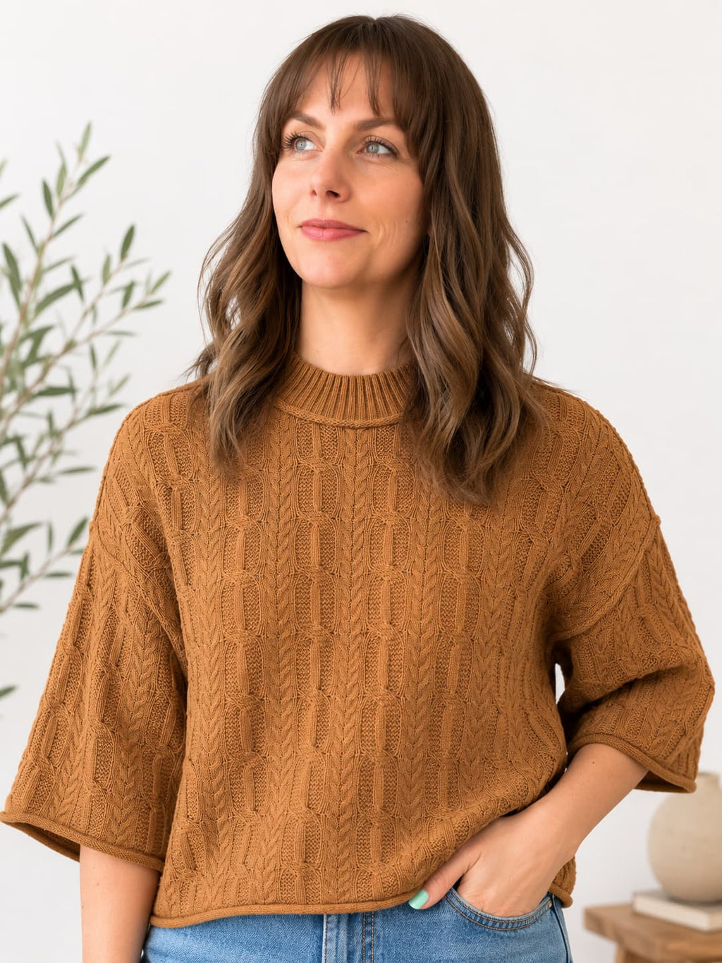 A woman wearing a textured, cable-knit short-sleeve sweater in a warm camel brown color, paired with blue jeans, standing against a clean white wall with a plant in the background.