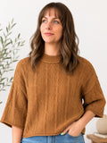A woman wearing a textured, cable-knit short-sleeve sweater in a warm camel brown color, paired with blue jeans, standing against a clean white wall with a plant in the background.